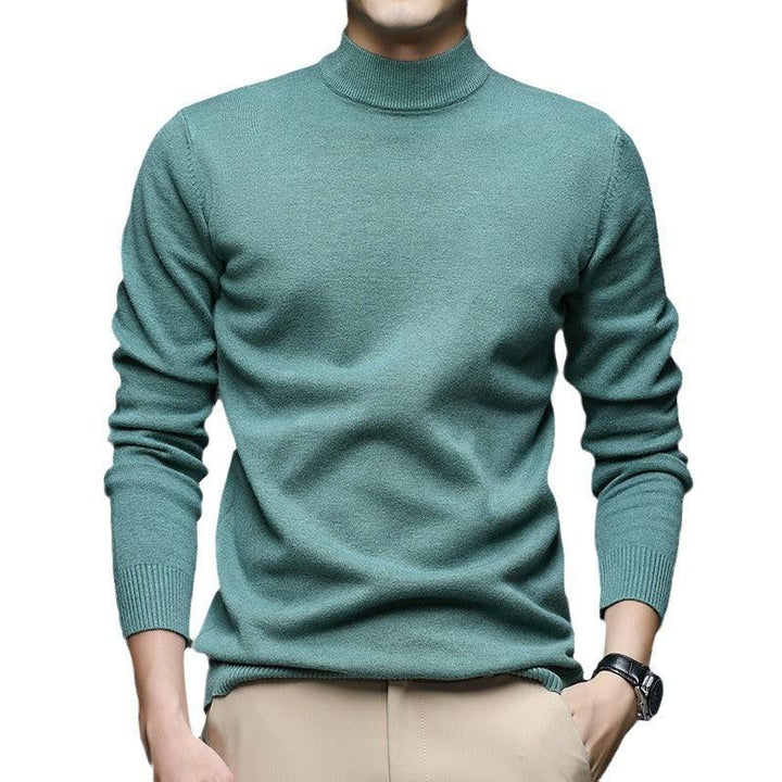 Man wearing teal turtleneck sweater and beige pants with watch on wrist against white background