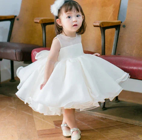 Toddler girl wearing white sleeveless party dress and headband indoors on wooden floor