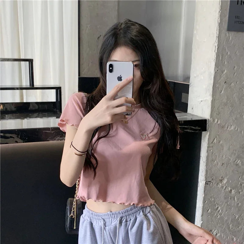 Young woman in a pink cropped top and gray sweatpants taking a mirror selfie with an iPhone in a modern indoor setting