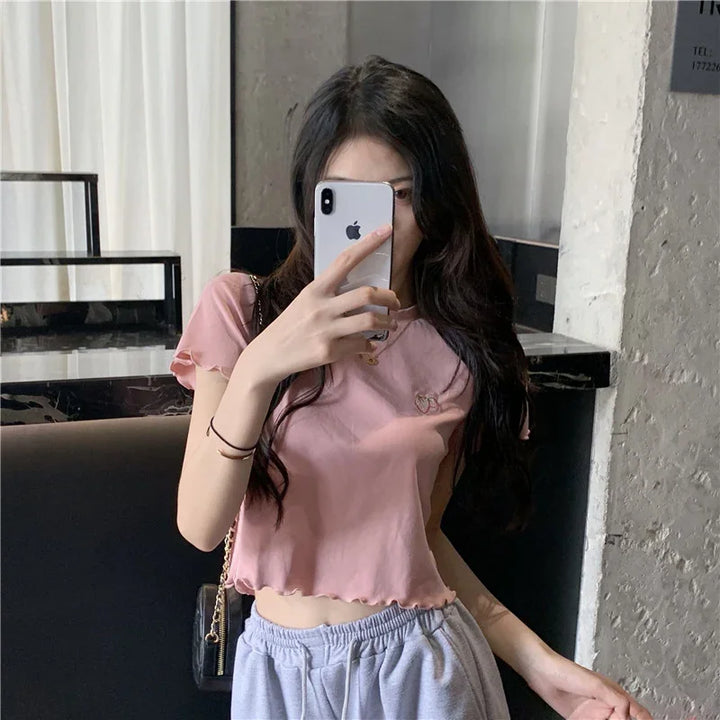 Young woman in a pink cropped top and gray sweatpants taking a mirror selfie with an iPhone in a modern indoor setting