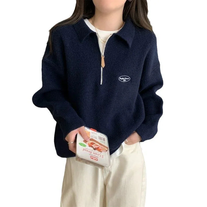 Woman wearing navy blue half-zip sweater with white logo and beige pants, holding packaged food