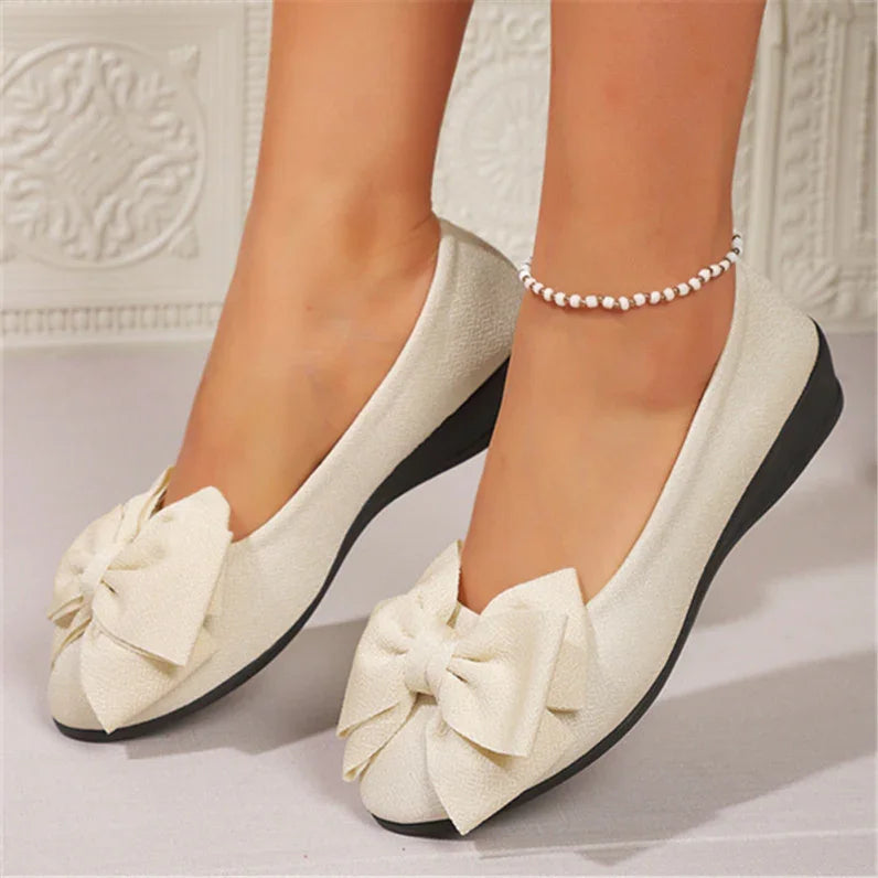 Cream-colored women's ballet flats with large fabric bows and black soles, worn with a beaded anklet