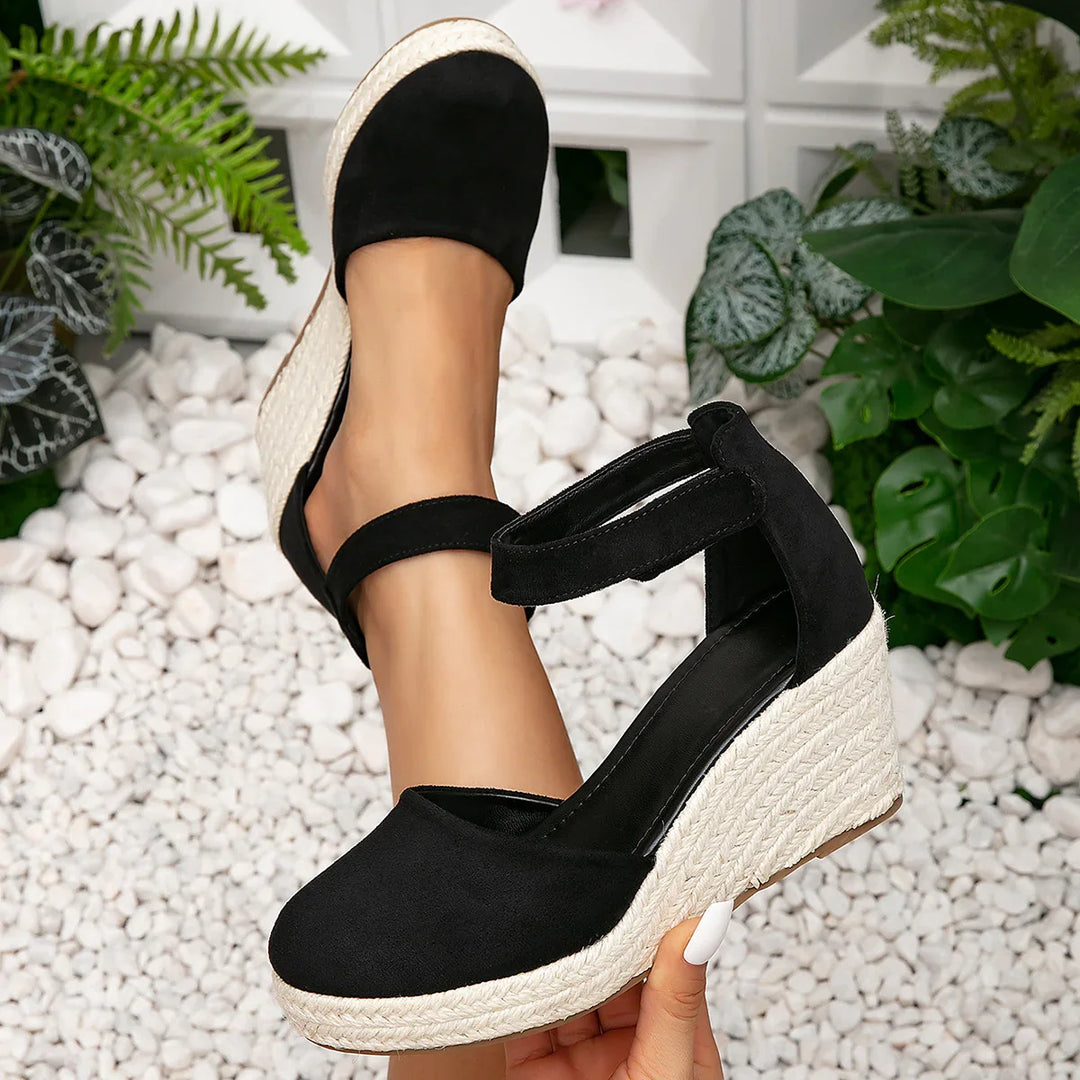 Black espadrille wedge shoes with ankle straps on feet over white pebbles and green plants background