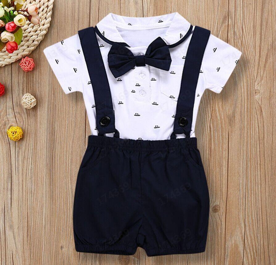 Baby boy formal outfit with white short-sleeve shirt, navy suspenders, bow tie, and shorts on wood background