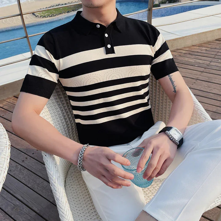 Man wearing black and white striped polo shirt and white pants sitting on white wicker chair near pool