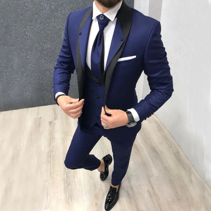 Man in a tailored navy blue three-piece suit with black lapels, white shirt, navy tie, and black dress shoes standing on light wood floor