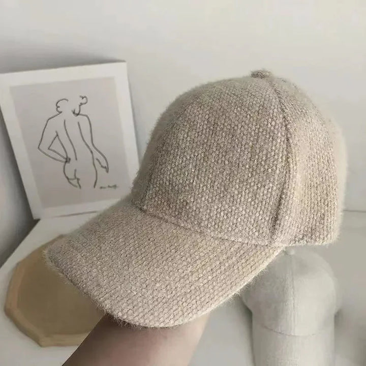 Close-up of beige textured wool baseball cap held indoors with abstract line art and another cap in background