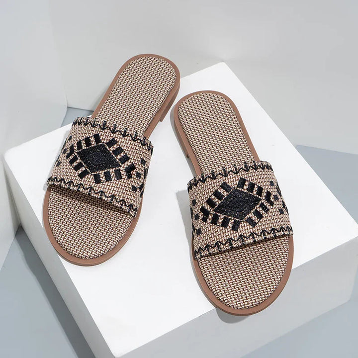 Brown woven slide sandals with black geometric embroidery on white display block