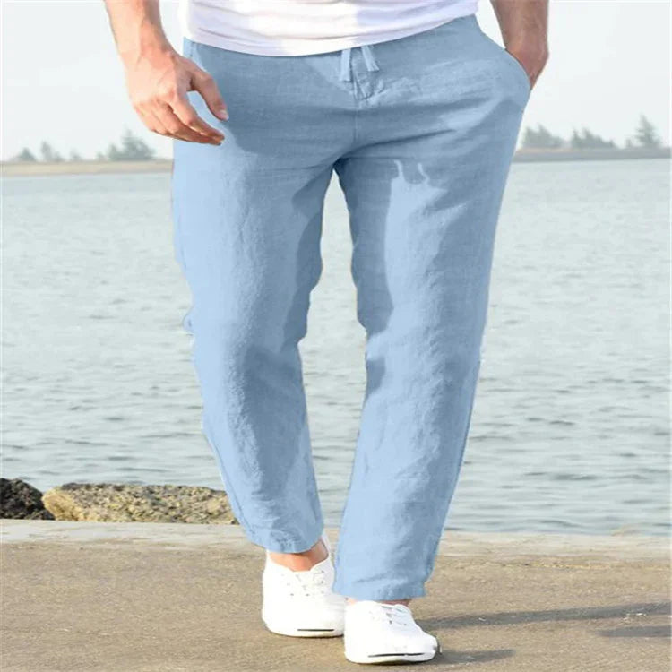 Man wearing light blue casual linen pants and white sneakers walking by the water on a sunny day