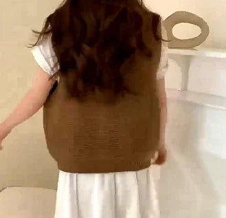 Woman wearing brown knitted vest over white dress with long wavy hair in minimal indoor setting