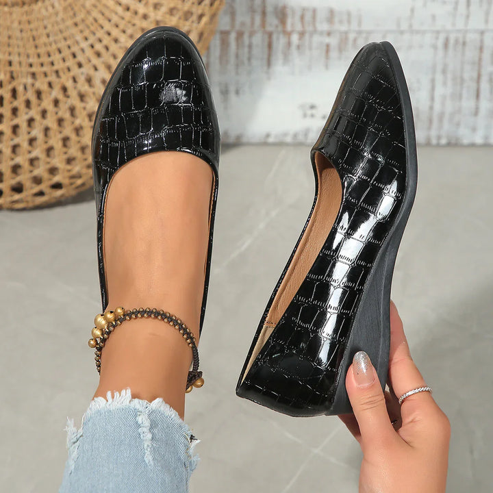Glossy black croc-patterned women's flats, one worn and one held showing almond toe design