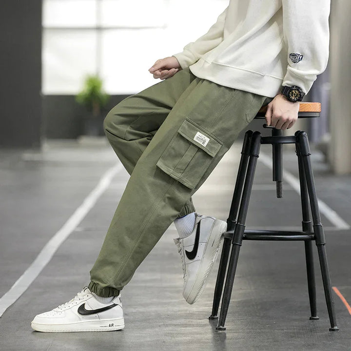 Person wearing olive green cargo pants with elastic cuffs and white Nike Air sneakers sitting on a black stool indoors