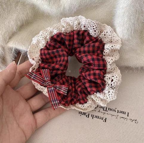 Hand holding a red and black plaid scrunchie with white lace trim and a matching bow on a beige fabric background