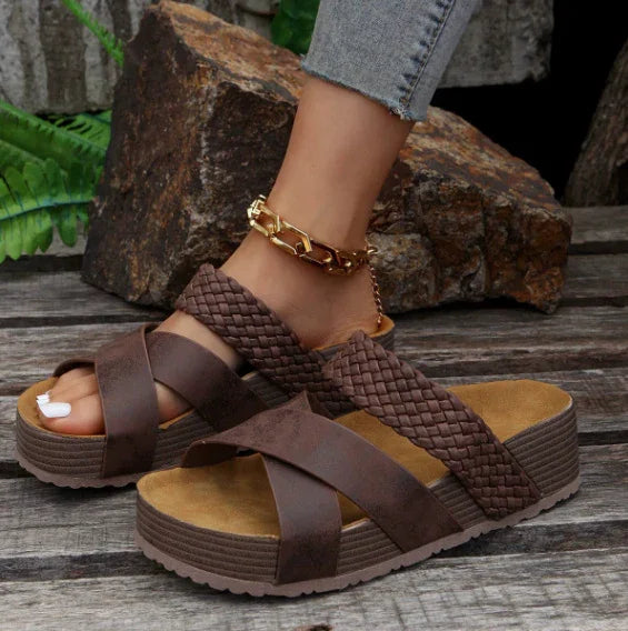 Brown wedge sandals with braided and smooth straps worn with gold anklet and light blue jeans on wooden surface