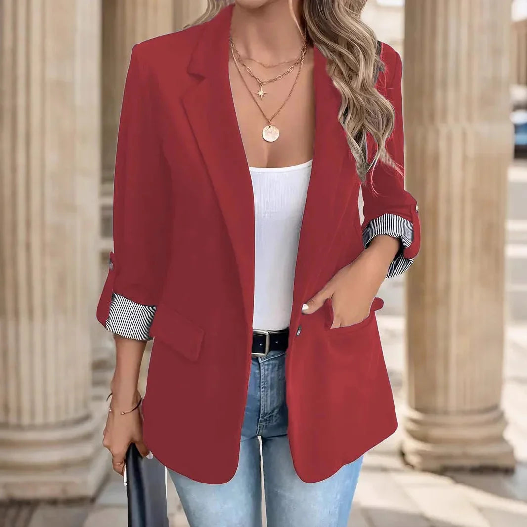 Woman wearing red blazer with rolled striped cuffs, white top, blue jeans, gold necklaces, and holding black clutch