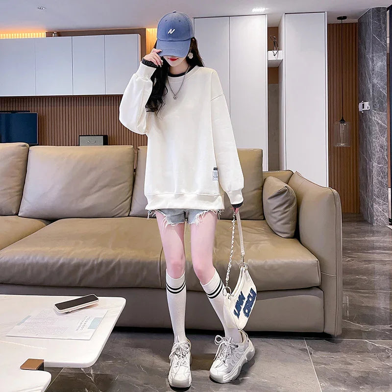 Woman in oversized white sweatshirt, denim shorts, knee-high socks, and sneakers indoors by beige sofa