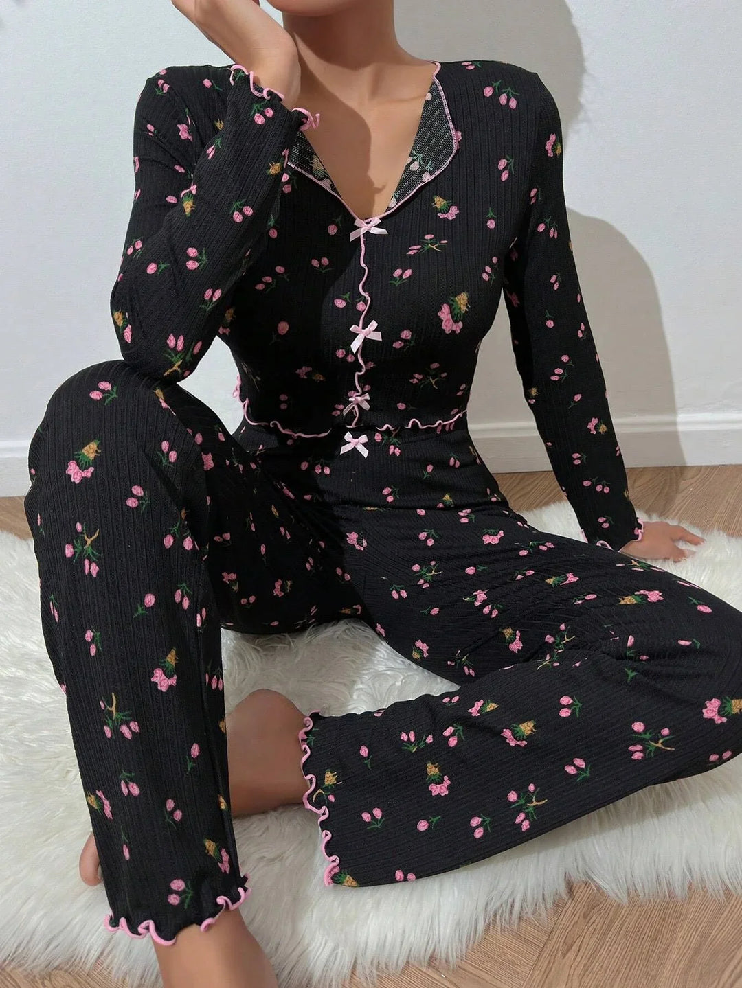 Woman wearing black floral ribbed knit pajama set with pink ruffle trim, sitting on white fur rug