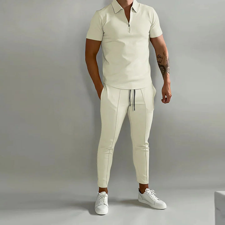 Man wearing beige short-sleeve zip polo shirt and matching jogger pants with white sneakers against gray background