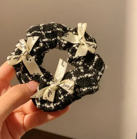 Hand holding a black and white textured fabric hair scrunchie with three cream ribbons tied in bows
