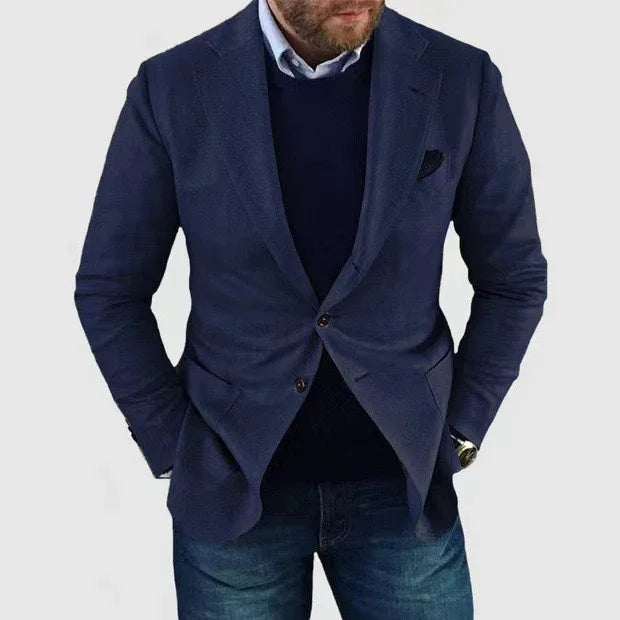 Man wearing a navy blue blazer with a black sweater, light blue dress shirt, and blue jeans