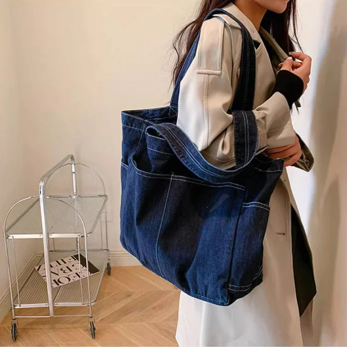 Woman wearing beige trench coat holding large dark blue denim tote bag indoors with metal cart in background