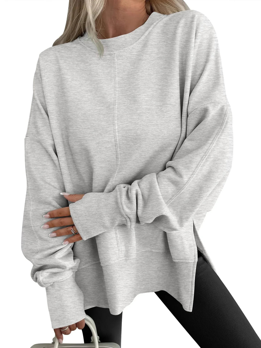 Woman wearing oversized light gray sweatshirt with long sleeves and black leggings holding a white handbag