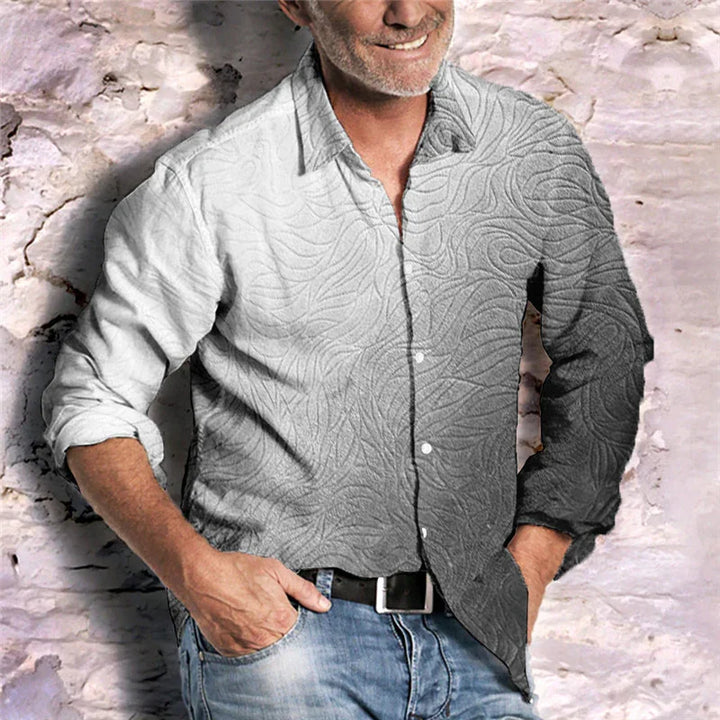 Smiling man in casual shirt with textured pattern and rolled-up sleeves, standing against rustic brick wall