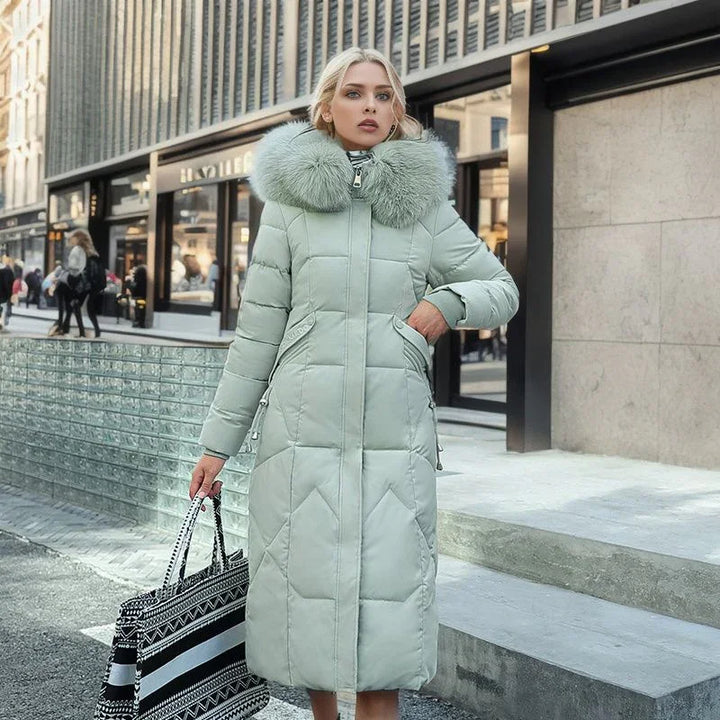 Woman wearing long light green padded winter coat with large fur collar holding black and white tote bag on city street