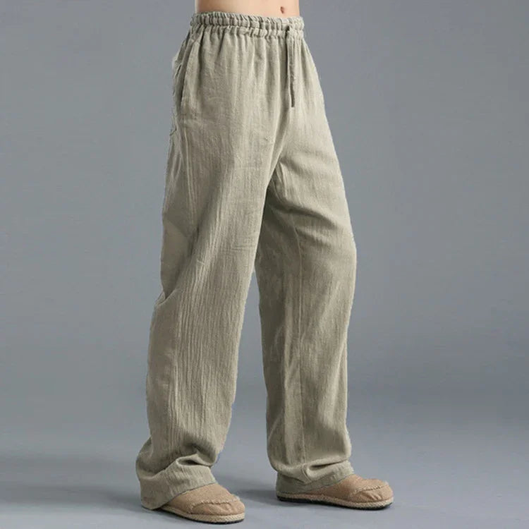 Person wearing loose beige linen pants with elastic waistband and slip-on shoes on gray background