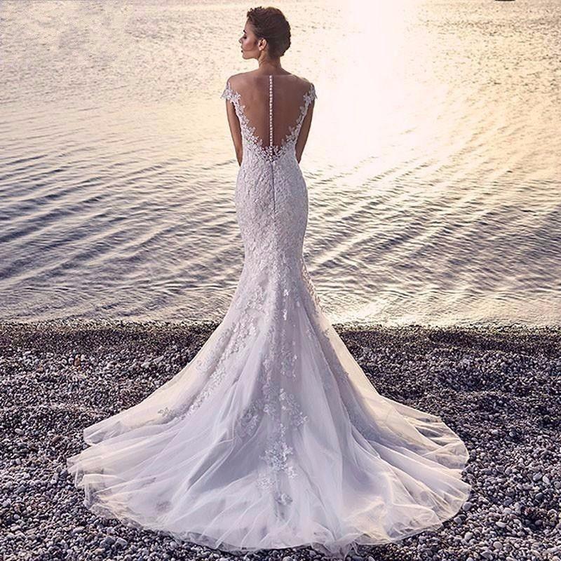 Bride in a lace mermaid wedding dress with sheer back standing on rocky shore by calm water at sunset