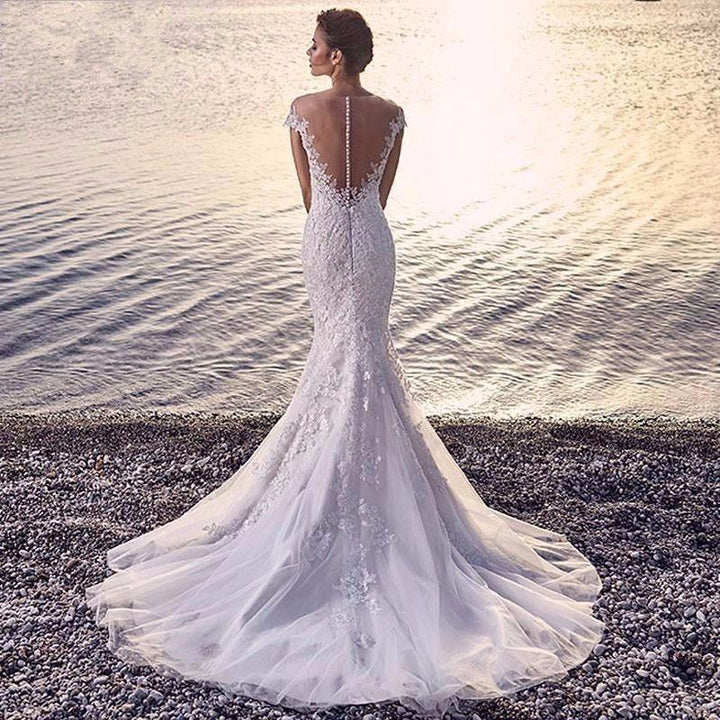 Bride in a lace mermaid wedding dress with sheer back standing on rocky shore by calm water at sunset