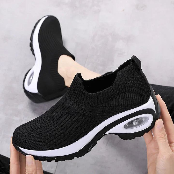 black slip-on knit sneakers with white air-cushioned sole held by hands on gray floor