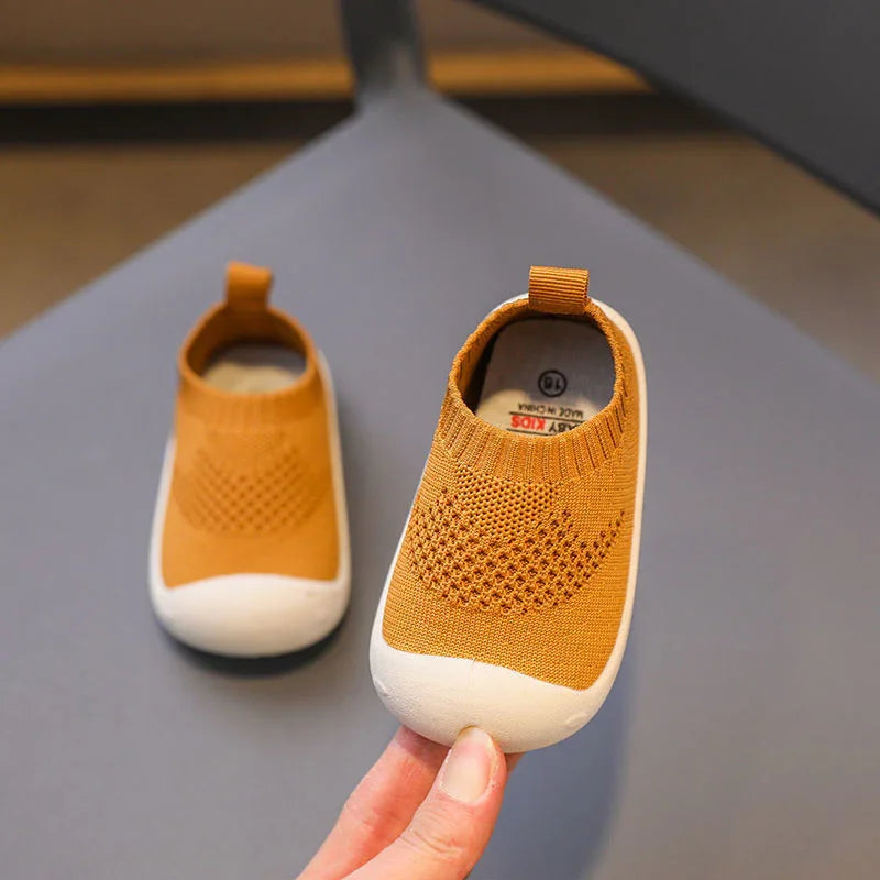 Pair of tan knit baby shoes with white rubber soles on gray surface, one held by fingers