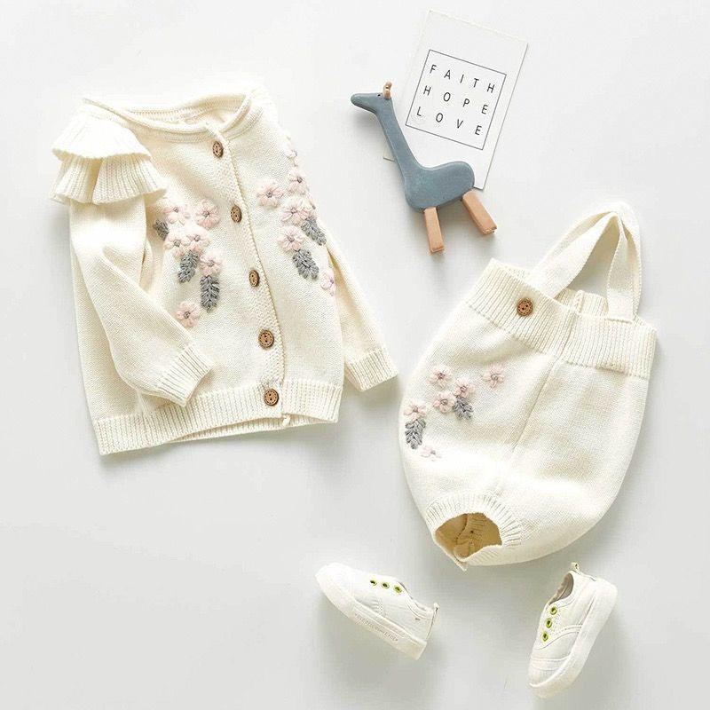Cream knit baby cardigan and romper with floral embroidery, white baby sneakers, blue wooden giraffe toy