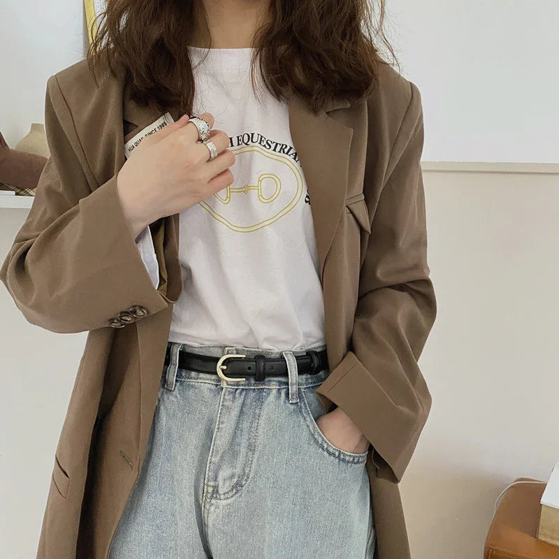 Woman wearing brown blazer, white graphic t-shirt, light blue jeans with black belt, and silver rings