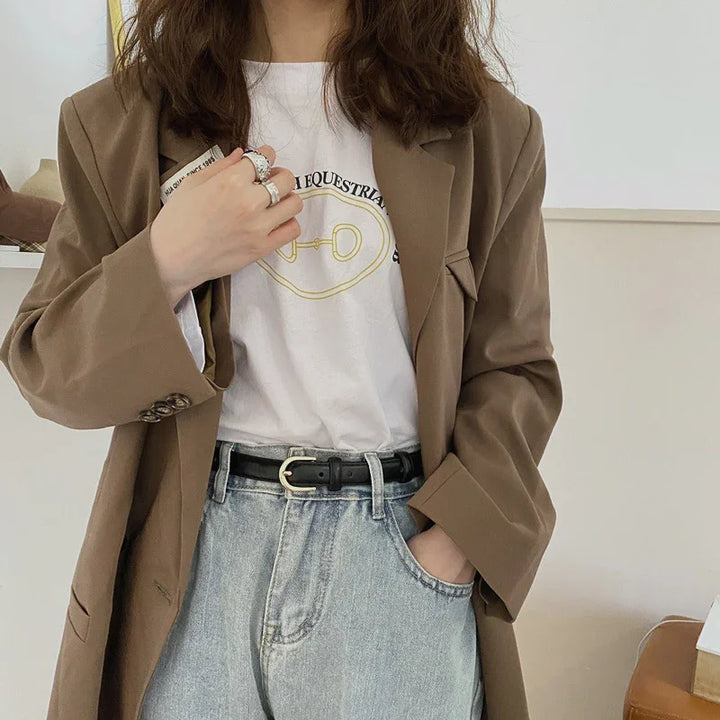 Woman wearing brown blazer, white graphic t-shirt, light blue jeans with black belt, and silver rings