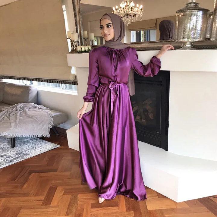 Woman in a purple satin long dress and brown hijab posing in a modern living room by a fireplace
