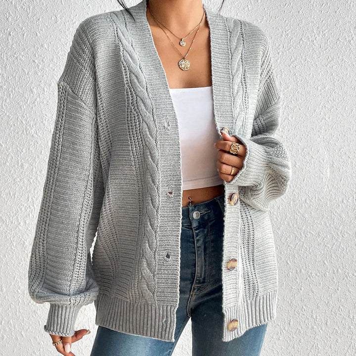 Woman wearing oversized gray cable knit cardigan with white crop top and blue jeans against textured white wall