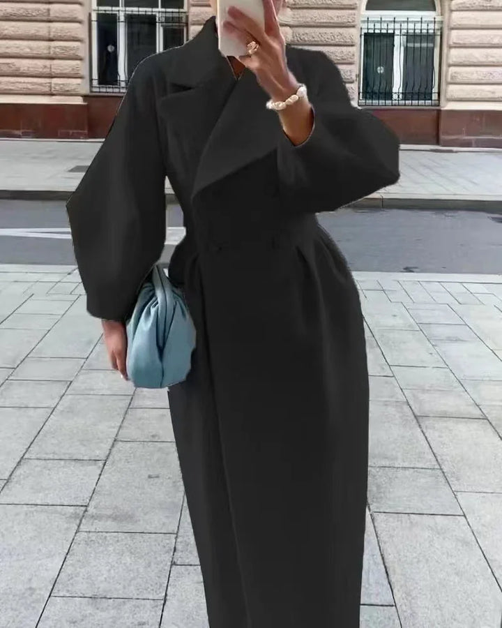Woman wearing a long black coat holding a blue handbag and taking a selfie on a city sidewalk
