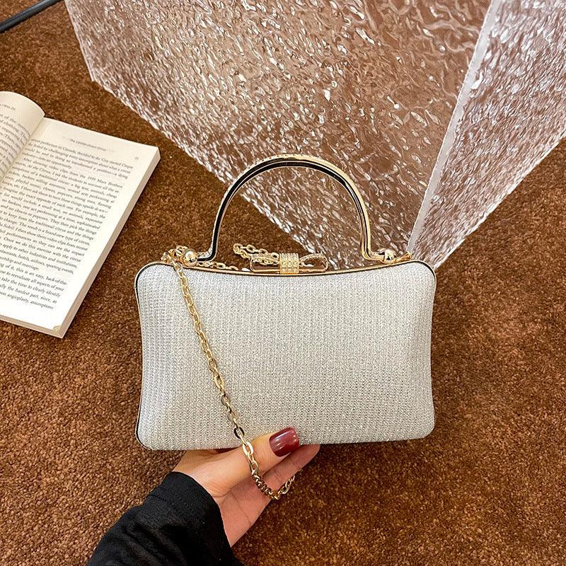 Elegant beige fabric clutch handbag with gold chain and metal handle held over brown carpet near open book