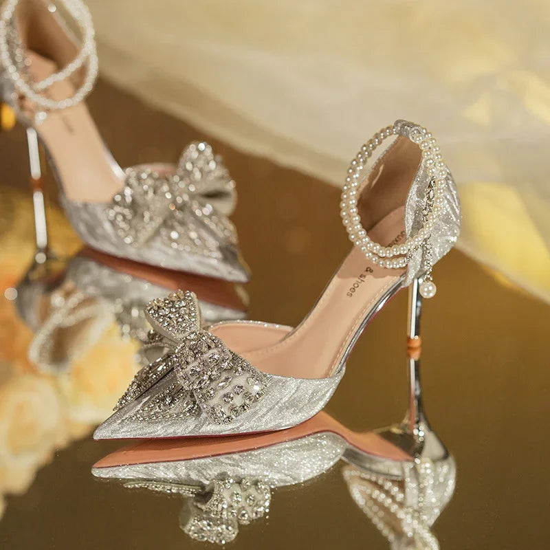 Elegant silver high-heeled shoes with crystal bow and pearl ankle strap on reflective surface