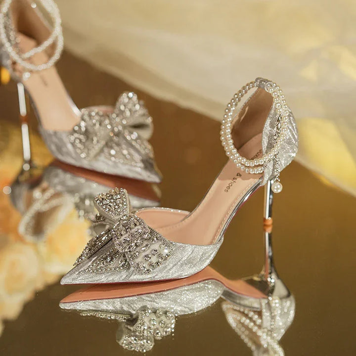 Elegant silver high-heeled shoes with crystal bow and pearl ankle strap on reflective surface