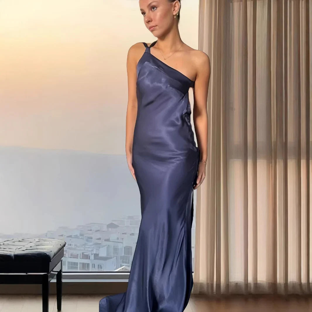 Woman wearing navy blue satin one-shoulder evening gown standing indoors near sheer curtains
