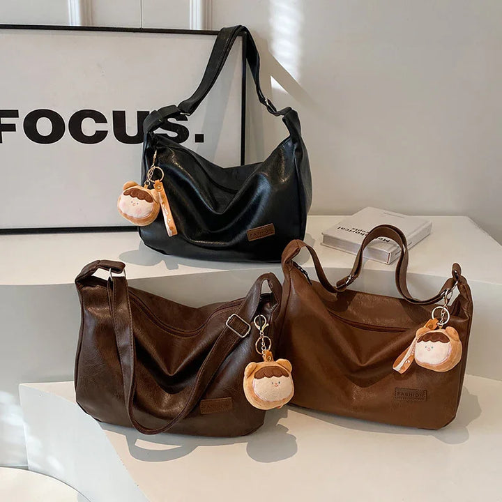 Three stylish leather shoulder handbags in black and brown with cute plush keychains on a white table