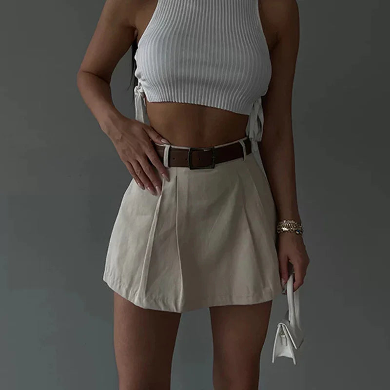 woman wearing white ribbed crop top and beige pleated mini skirt with brown belt holding small white handbag