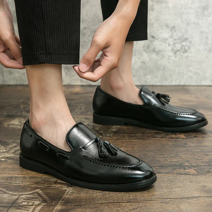 Black leather tassel loafers worn with cropped black trousers on wooden floor