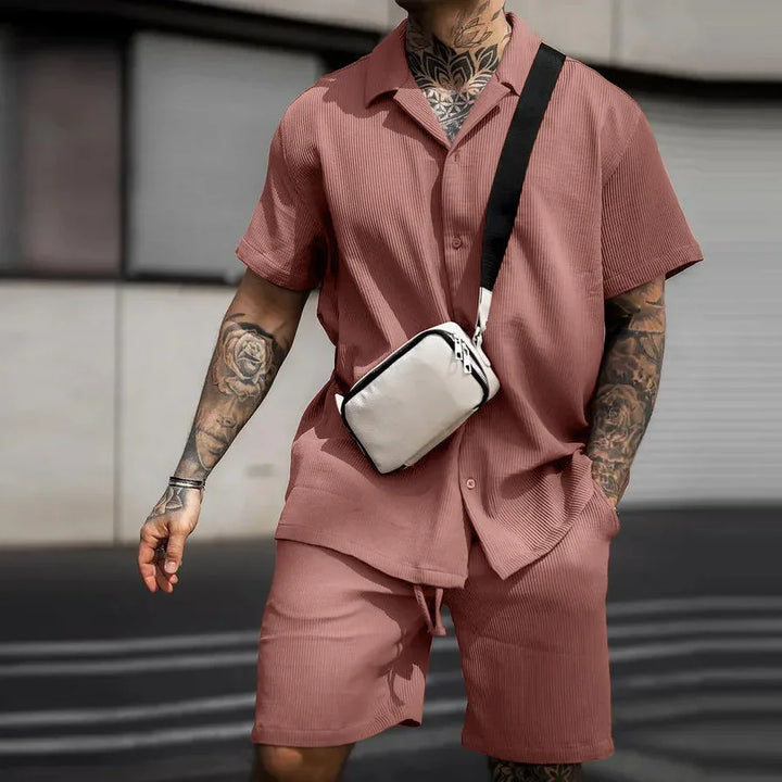 Man wearing dusty pink ribbed short-sleeve shirt and shorts with black tattooed arms and white crossbody bag