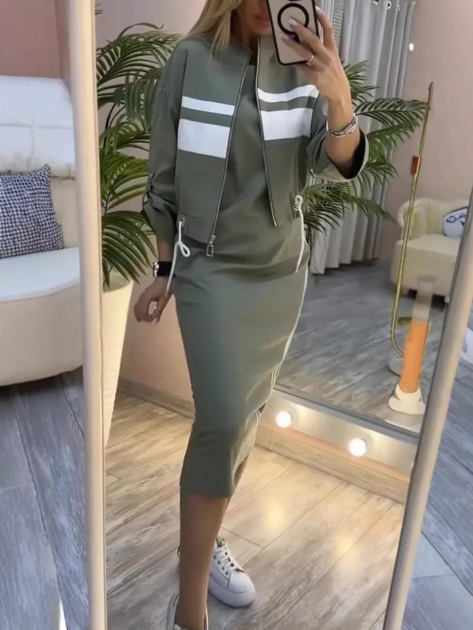 Woman wearing olive green midi dress and matching jacket with white stripes, posing in modern room mirror