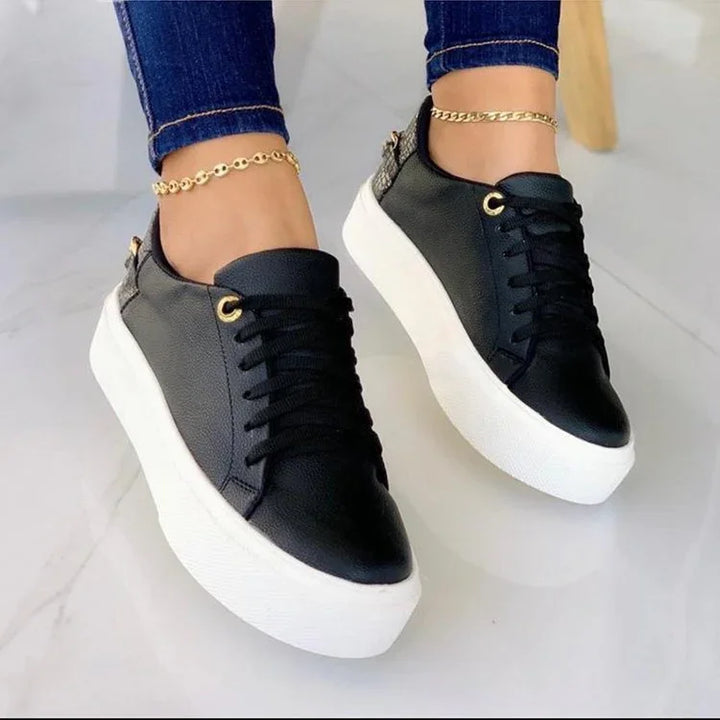 Black leather platform sneakers with white thick soles worn with blue jeans and gold anklets on a white floor