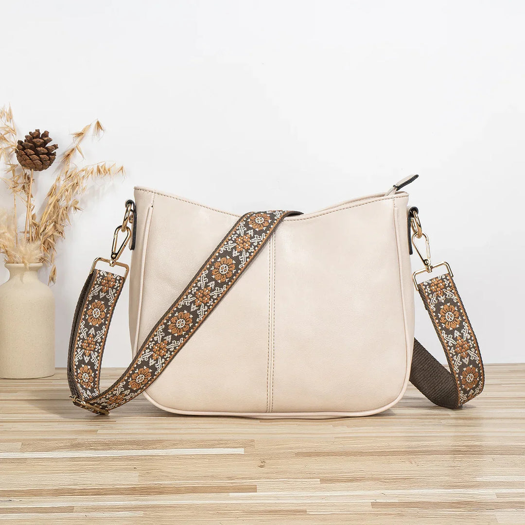Beige leather crossbody bag with brown embroidered floral strap on wooden surface